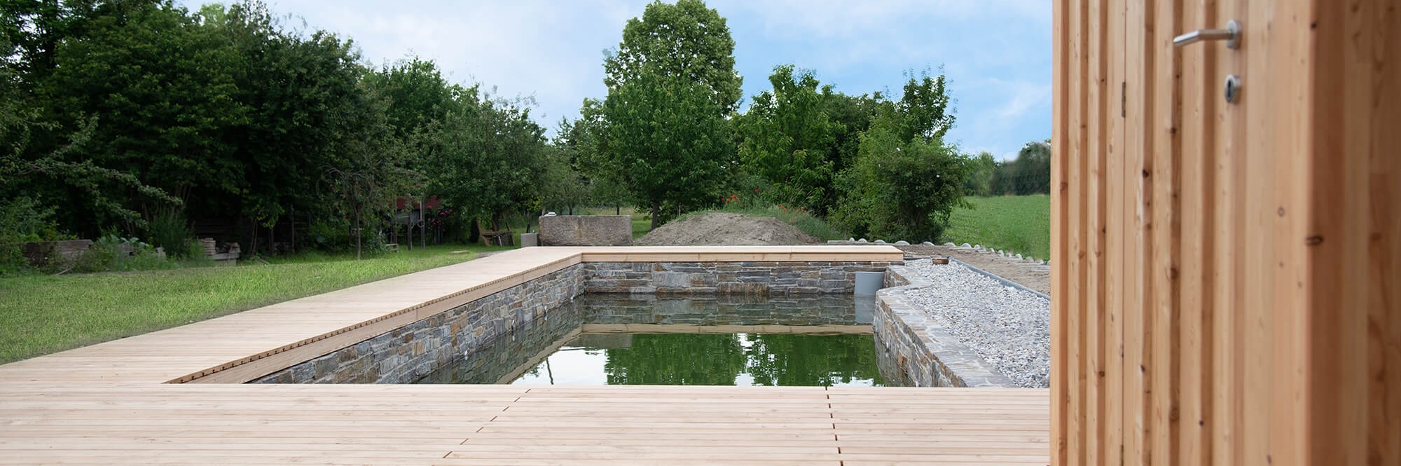 Holzterrasse mit integriertem Naturteich auf einer Wiese neben einer natürlichen grünen Hecke.