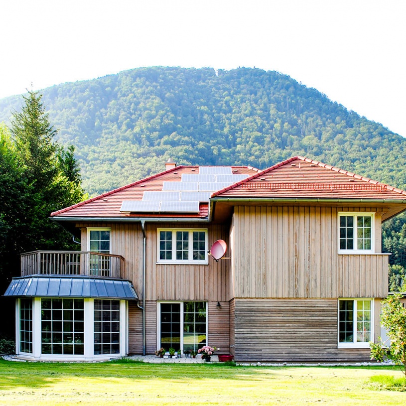 Ein Haus aus Holz hat ein rotes Dach und einen Garten mit einem kleinen Baum auf der rechten Seite