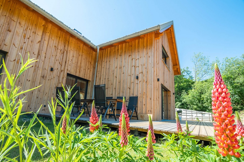 Ein Garten mit Blumen und ein Holzhaus mit Terrasse, auf dem ein schwarzer Tisch mit schwarzen Stühlen steht