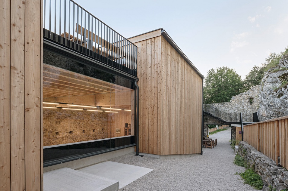 Große Glasfront inmitten der Holzfassade links. Rechts finden sich Burgmauern