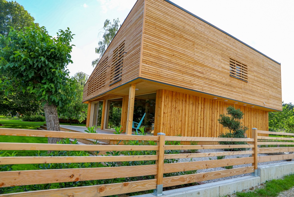 Hinter einem Holzzaun befindet sich ein zweistöckiges Wohnhaus aus Holz mit Glastüren.
