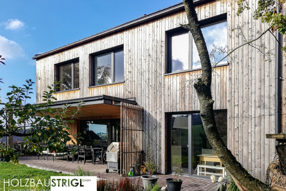 Ein zweistöckiges Haus aus ausgeblichenem Holz mit großen Fenstern und einem Garten, in dem ein kleiner Baum steht