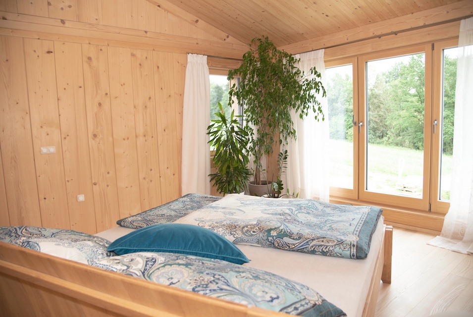 Mit Holz verkleidetes Gästezimmer. Zu sehen ist ein großes Bett im Vordergrund und Blick ins Grüne
