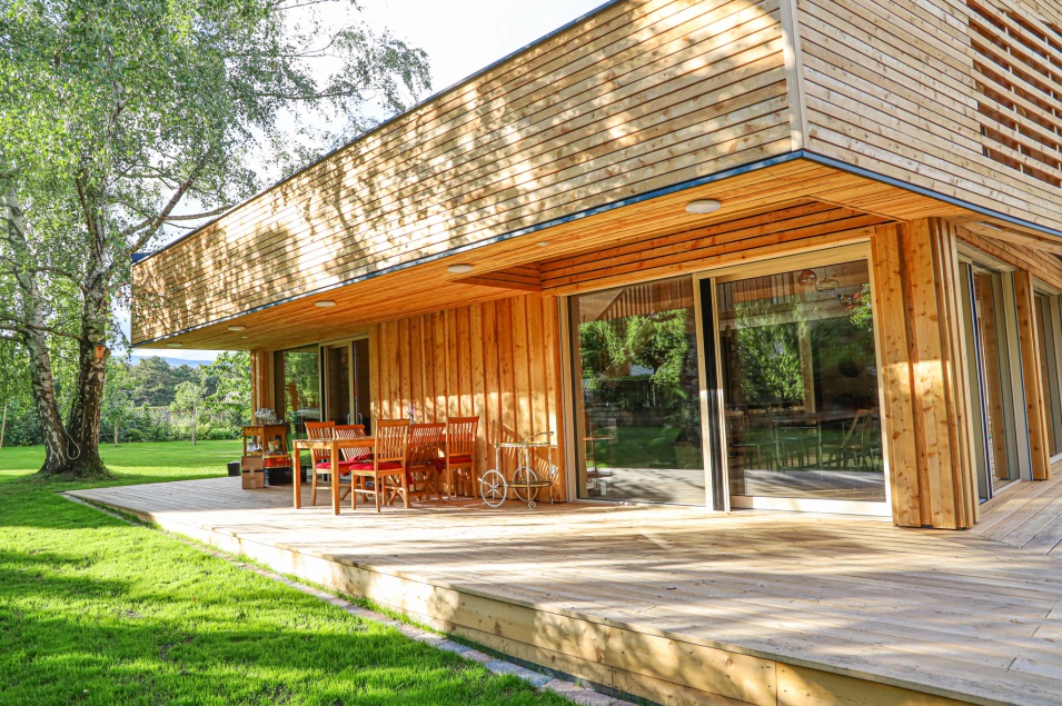 Ein zweistöckiges Holzhaus mit großzügiger Terrasse, auf der sich ein Tisch mit 6 Stühlen befindet