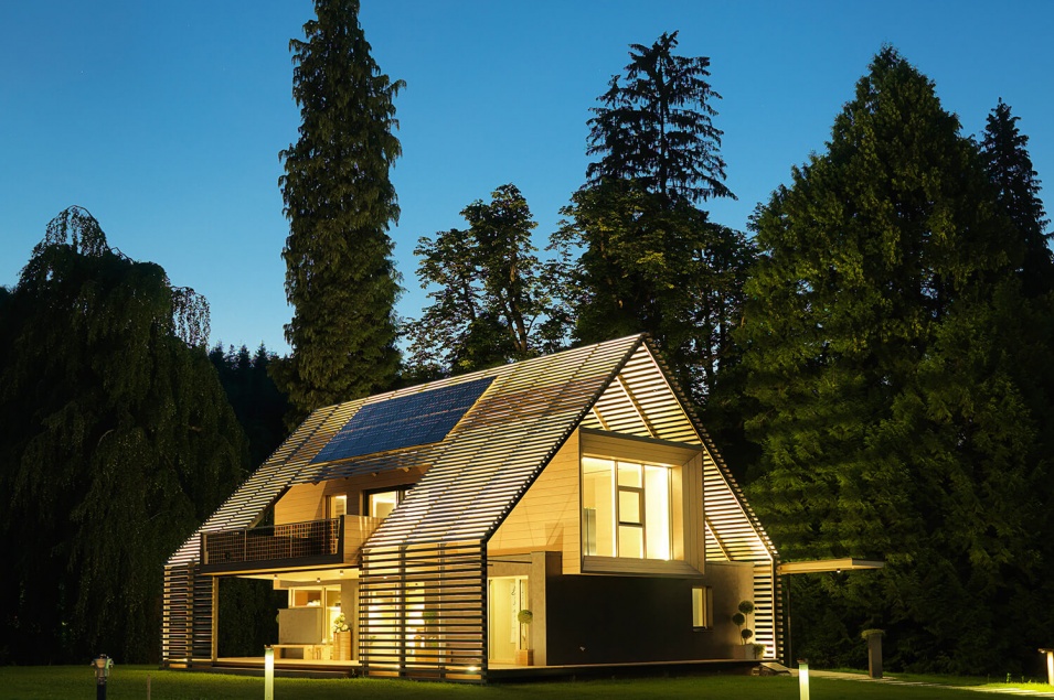 Ein stark beleuchtetes modernes Passivhaus aus Holz mit Satteldach und einem Wald dahinter