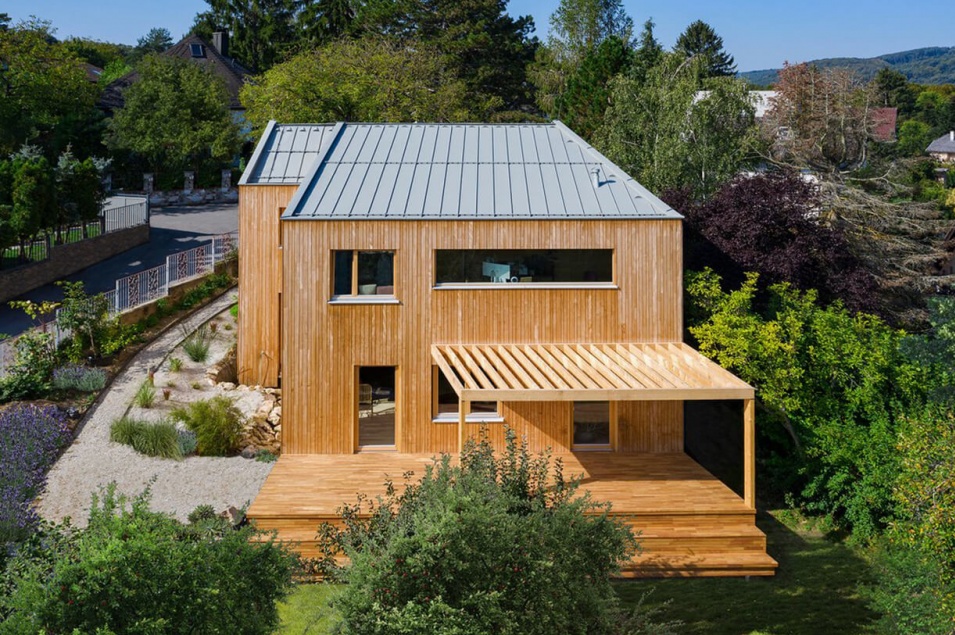 Ein Haus komplett aus Holz ist von vielen grünen Pflanzen umgeben und hat eine überdachte Terrasse.