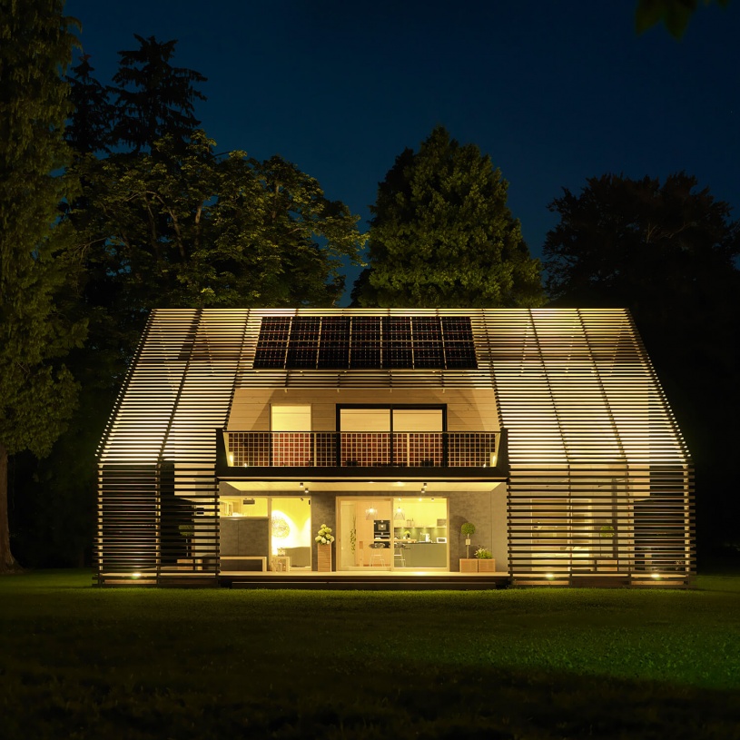 Ein stark beleuchtetes Passivhaus aus Holz mit Satteldach und Blick in die Küche vom Garten aus