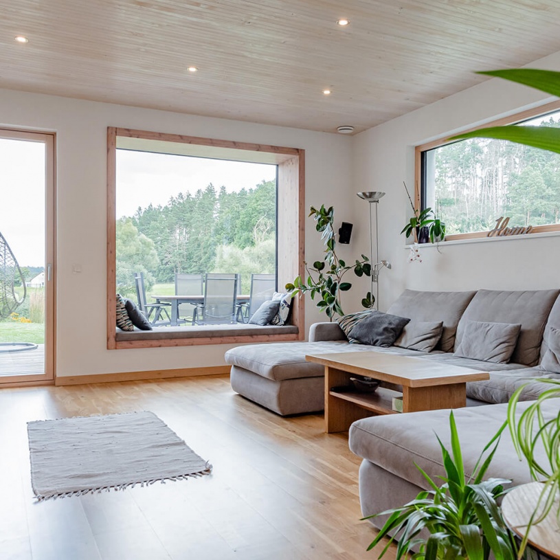Ein Wohnzimmer mit grauer Couch, großen Fenstern, einem grauen Teppich und hellem Holzboden