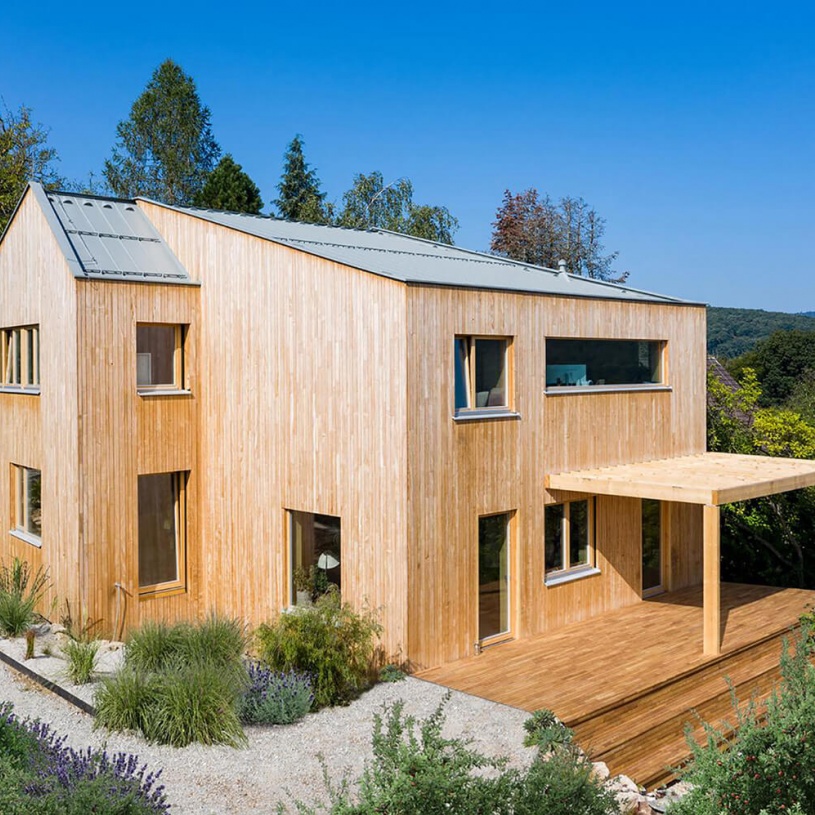 Ein Haus aus Holz mit vielen Fenster und Kieselsteinen im Vorgarten ist umgeben von vielen grünen Pflanzen.