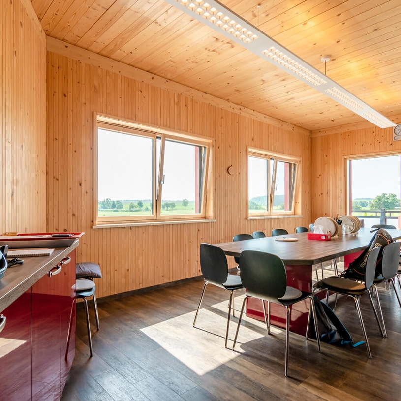 Mit hellem Holz verkleideter Aufenthaltsraum, großem dunklen Tisch mit einigen Sesseln rechts und roter Kommode links