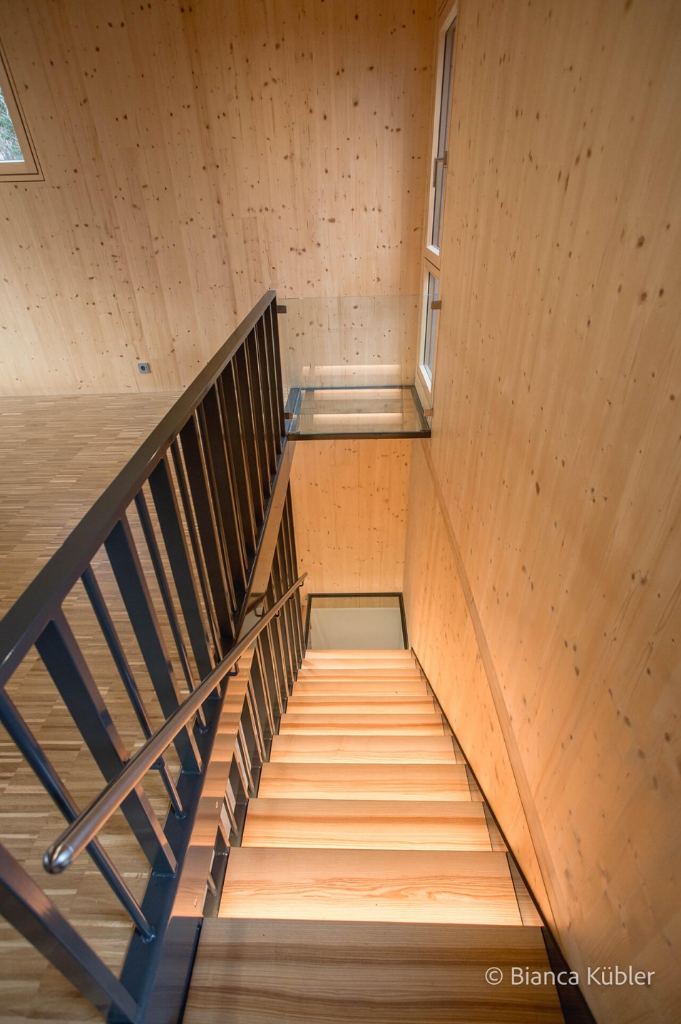 Blick von einer beleuchteten, hellen Holzstiege in den untern Stock mit grauem Geländer links und einer Holzwand rechts