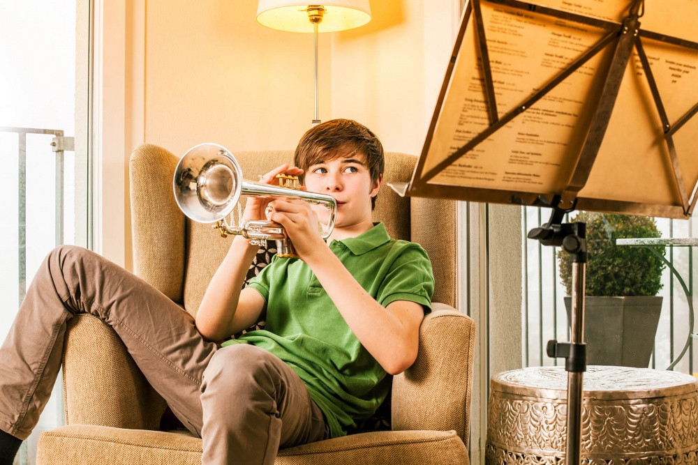 Ein Junge der in einem braunen Sofa sitzt spielt auf einer Trompete und blickt auf einen Notenständer im Vordergrund